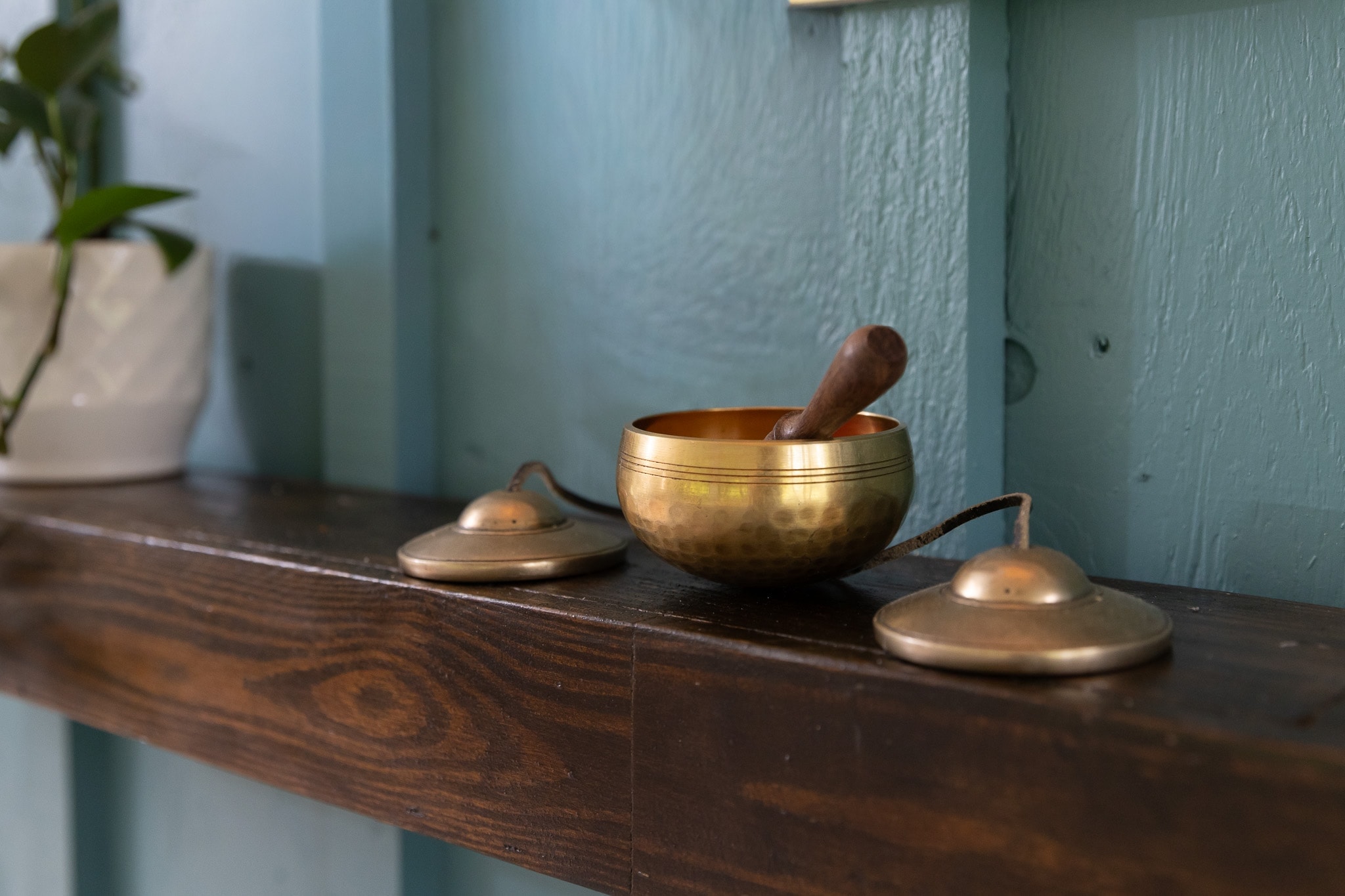 Brass singing bowl and tingsha cymbals resting on a wooden shelf against a teal wall